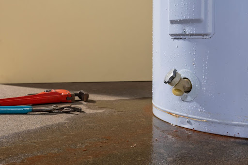 Old water heater leaking water on basement floor with plumbing tools sitting right next to it.